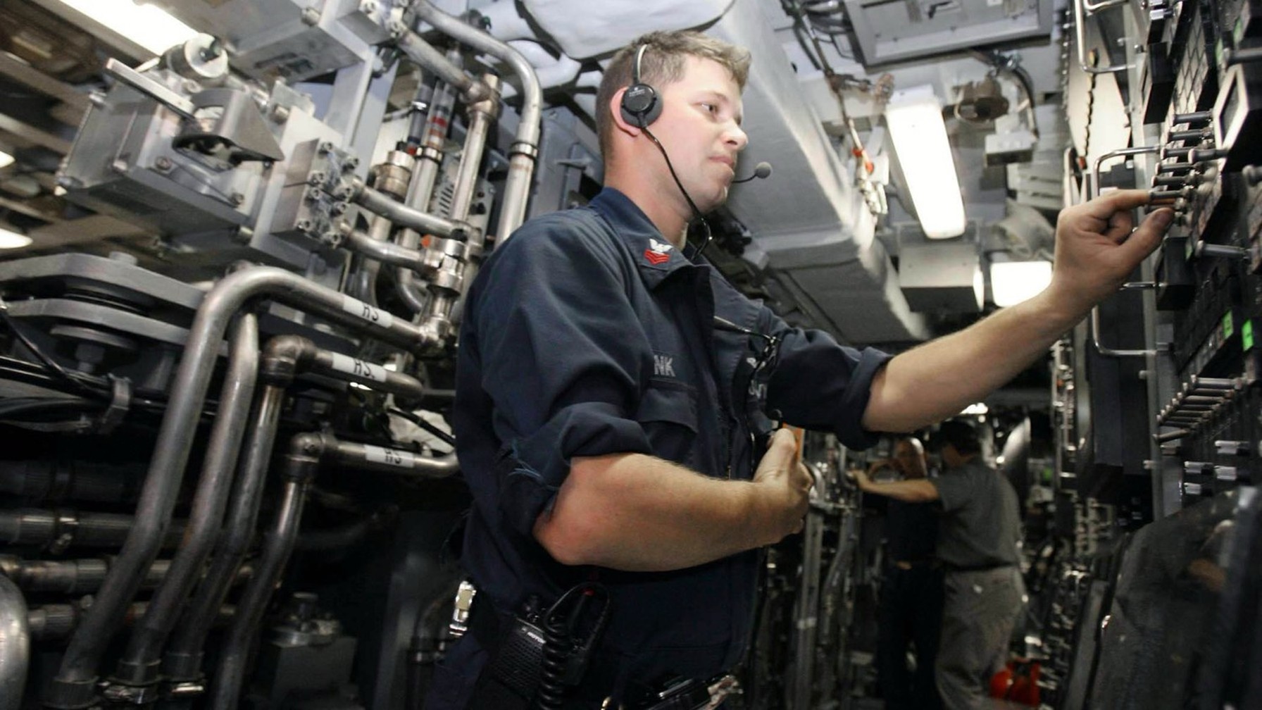 A U.S. Navy sailor in a headset reaches for control levers among pipes and valves inside a submarine